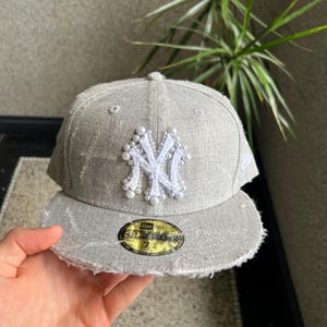 Könnte beinhalten: Graue Baseballkappe mit ausgefranstem Rand und einem weiß und perlenbesetzten New York Yankees-Logo. Die Kappe hat einen gebogenen Schirm und einen 59FIFTY-Aufkleber.