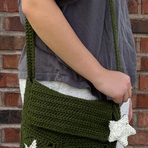May include: Olive green crochet shoulder bag with white star appliqués. The bag has a flap closure and a long strap. The bag is made of a textured yarn and has a handmade appearance. The bag is a stylish accessory.
