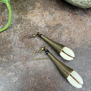 May include: A pair of handcrafted earrings featuring a white shell pendant encased in a spiral of brass-colored wire. The earrings have a hook closure and a small black bead detail. The design is set against a textured, brown surface.