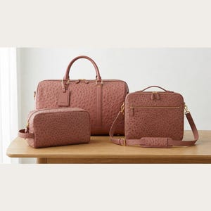 May include: A set of three matching dusty rose-colored bags with a textured finish. The set includes a large duffel bag, a briefcase with a shoulder strap, and a smaller toiletry bag. All bags have gold-toned hardware and are displayed on a wooden table.