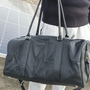 May include: A black duffel bag with a snakeskin texture. The bag is held by a person. It features two long handles and a detachable shoulder strap. Silver-coloured hardware accents the bag's design. The bag is made of a durable material.