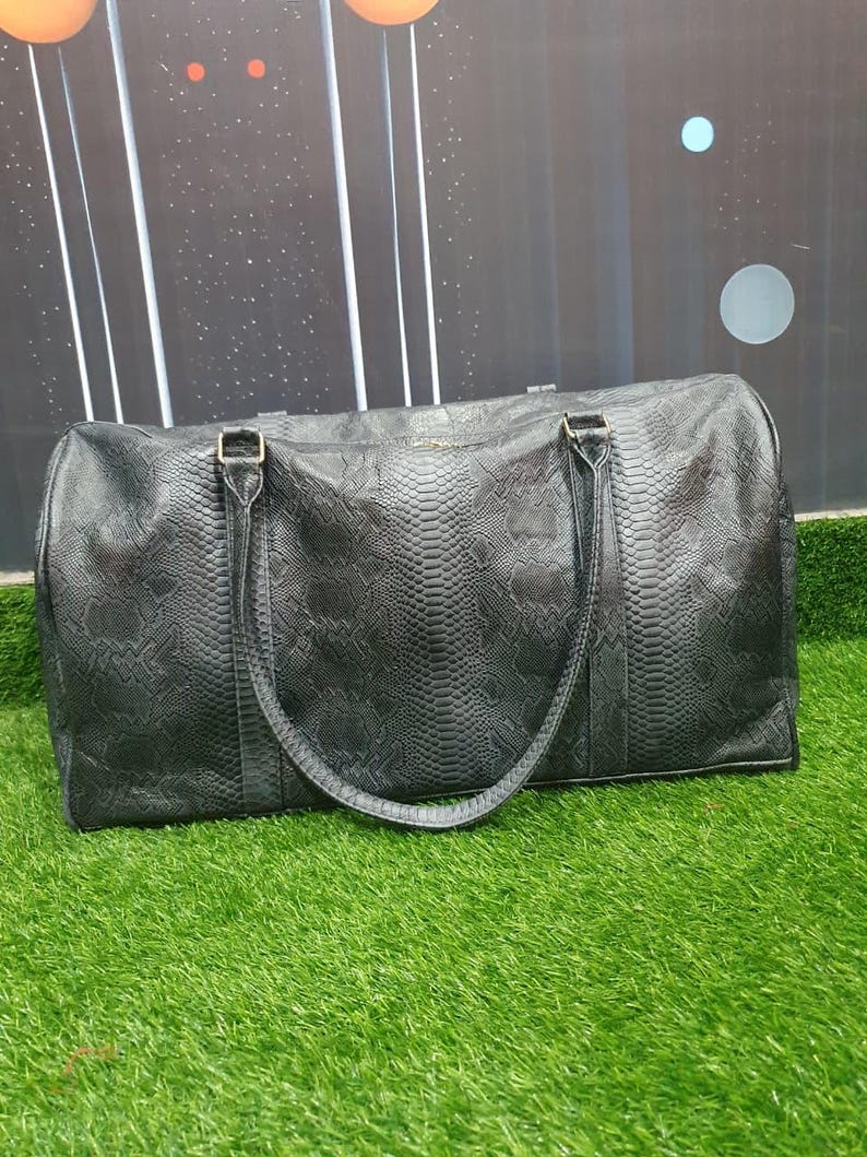 Pode incluir: Uma mala de viagem preta com um padr&atilde;o texturizado de pele de cobra em toda a sua superf&iacute;cie. A mala possui duas al&ccedil;as pretas longas e a condizer fixadas no topo, um fecho de correr horizontal e uma forma retangular com cantos arredondados.