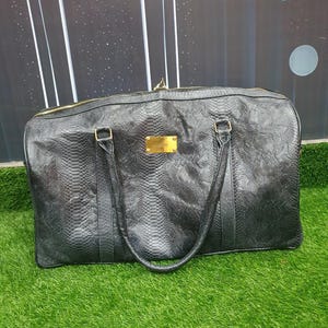 Pode incluir: Uma mala de viagem preta com um padr&atilde;o texturizado de pele de cobra, duas al&ccedil;as superiores e uma placa met&aacute;lica retangular dourada centrada na parte frontal. A mala inclui um fecho de correr dourado na borda superior e ferragens douradas.