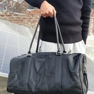 Op de afbeelding: Zwarte duffeltas met een textuur van slangenleer. De tas heeft twee handvatten aan de bovenkant en een afneembare schouderriem. De tas wordt vastgehouden door een persoon die een zwarte trui en een grijze joggingbroek draagt.