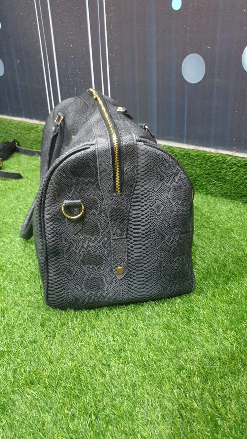 Pode incluir: Uma mala de viagem cinzento-escura com um padr&atilde;o texturizado de pele de cobra. A mala inclui um fecho de correr dourado na parte superior, um anel em D dourado na lateral e um pequeno detalhe de rebite dourado junto ao fecho.