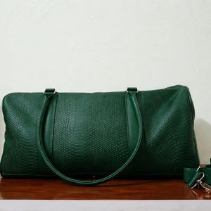May include: A dark green faux leather duffel bag with a textured, snake-skin pattern. The bag has two top handles and a detachable shoulder strap. The bag is sitting on a wooden surface.