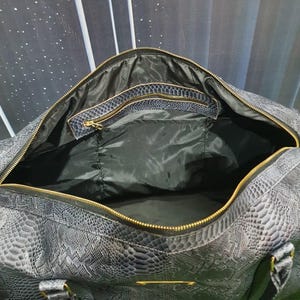 Pode incluir: Uma mala de viagem cinzento-escura com padr&atilde;o texturizado de pele de cobra, apresentando um fecho de correr dourado, forro interior preto e um bolso interior com fecho e acabamento a condizer, incluindo pegas resistentes com ferragens douradas.