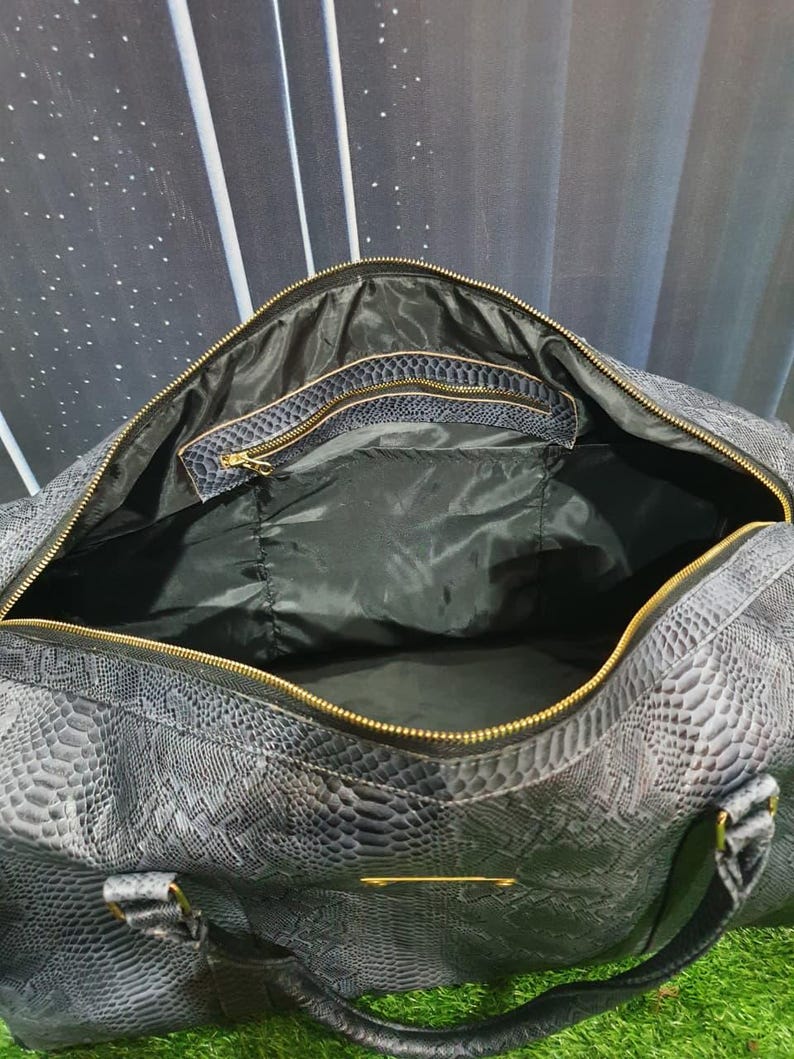 Pode incluir: Uma mala de viagem cinzento-escura com padr&atilde;o texturizado de pele de cobra, apresentando um fecho de correr dourado, forro interior preto e um bolso interior com fecho e acabamento a condizer, incluindo pegas resistentes com ferragens douradas.