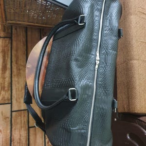 May include: Black duffel bag with a textured, snake-skin-like pattern. The bag has a silver zipper, a top handle, and a shoulder strap. The bag is rectangular and appears to be made of leather.