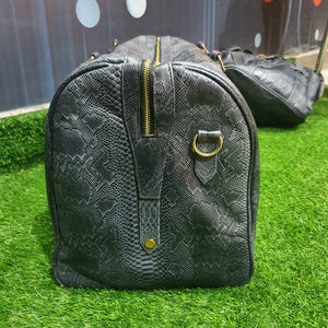 Pode incluir: Uma mala de viagem cinzento-escura com um padr&atilde;o texturizado de pele de cobra. A mala apresenta um fecho central preto com ferragens em tom dourado, incluindo um anel lateral circular e um pequeno rebite dourado na base do fecho.