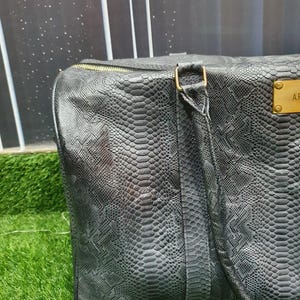 Pode incluir: Uma mala de viagem cinzento-escura com um padr&atilde;o texturizado de pele de cobra, apresentando um fecho de correr dourado e uma placa met&aacute;lica retangular dourada na frente com o texto gravado A.F. A mala inclui uma al&ccedil;a a condizer com detalhes dourados.