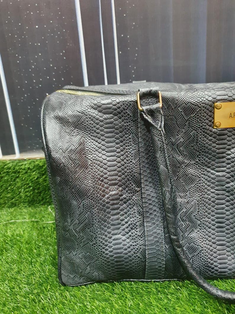 Pode incluir: Uma mala de viagem cinzento-escura com um padr&atilde;o texturizado de pele de cobra, apresentando um fecho de correr dourado e uma placa met&aacute;lica retangular dourada na frente com o texto gravado A.F. A mala inclui uma al&ccedil;a a condizer com detalhes dourados.