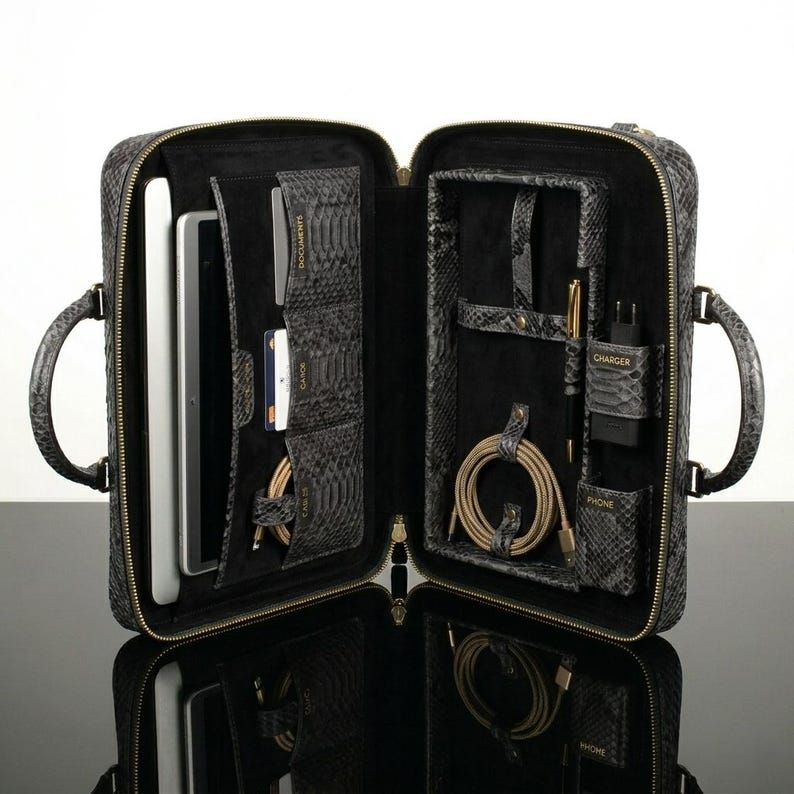 Pode incluir: Maleta cinza aberta com padr&atilde;o de pele de cobra e z&iacute;per dourado. O interior revela compartimentos para um tablet, cart&otilde;es, cabos, uma caneta e um carregador. A maleta tem duas al&ccedil;as e uma superf&iacute;cie refletora.