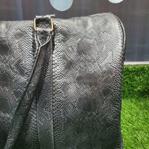 Pode incluir: Uma mala de viagem preta com um padr&atilde;o texturizado de pele de cobra em toda a superf&iacute;cie. A mala inclui um detalhe de al&ccedil;a vertical com uma fivela retangular de cor dourada met&aacute;lica que liga a pega ao corpo principal.