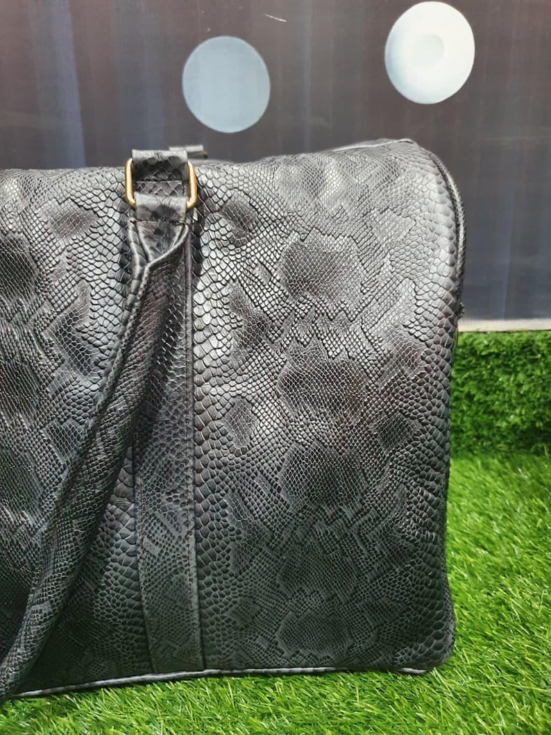 Pode incluir: Uma mala de viagem preta com um padr&atilde;o texturizado de pele de cobra em toda a superf&iacute;cie. A mala inclui um detalhe de al&ccedil;a vertical com uma fivela retangular de cor dourada met&aacute;lica que liga a pega ao corpo principal.