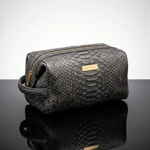 Pode incluir: Uma bolsa de viagem cinza com textura de pele de cobra, z&iacute;per e al&ccedil;a dourados. A bolsa tem formato retangular com uma pequena etiqueta dourada que diz "Forge Leather Co."