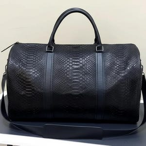 Op de afbeelding: Een zwarte duffeltas met een slangenleerpatroon. De tas heeft een handvat aan de bovenkant en een schouderriem. De tas is rechthoekig en heeft een ritssluiting. De tas is gemaakt van leer en heeft een strak, modern design.