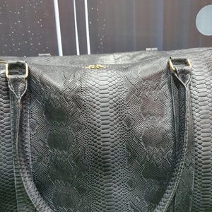 Pode incluir: Uma mala de viagem preta com um padr&atilde;o texturizado de pele de cobra em todo o exterior. A mala inclui duas al&ccedil;as resistentes presas com ferragens met&aacute;licas em tom dourado e um fecho de correr central ao longo da borda superior da mala.