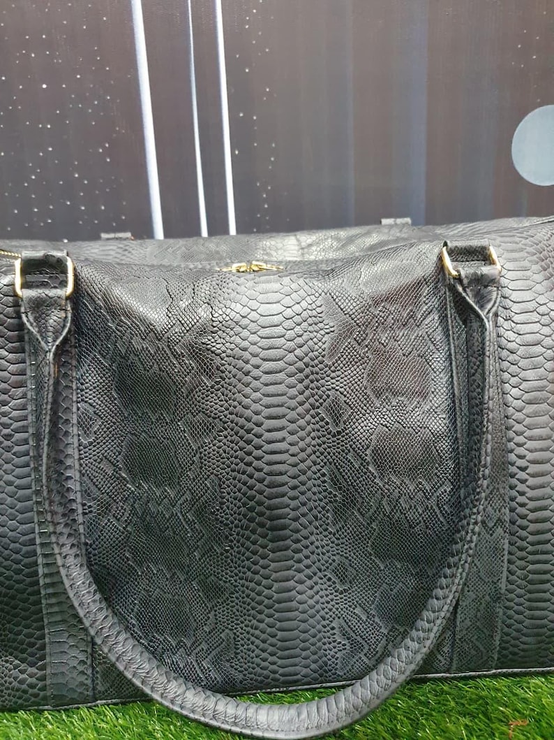 Pode incluir: Uma mala de viagem preta com um padr&atilde;o texturizado de pele de cobra em todo o exterior. A mala inclui duas al&ccedil;as resistentes presas com ferragens met&aacute;licas em tom dourado e um fecho de correr central ao longo da borda superior da mala.