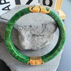 Könnte beinhalten: Ein leuchtend grünes Jade-Armreif-Armband mit goldfarbenen Verschlüssen. Der Jade hat ein gesprenkeltes Aussehen, und die goldenen Verschlüsse weisen dekorative Designs auf. Das Armband wird auf einer strukturierten grauen Oberfläche präsentiert.