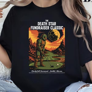 May include: Black t-shirt featuring a colorful graphic of a golfer in armor on a golf course with a Death Star in the background. The text reads "THE DEATH STAR FUNDRAISER CLASSIC". The shirt also has additional text at the bottom.