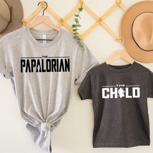 May include: Two gray t-shirts with black text. The larger shirt says "The Papalorian" and the smaller shirt says "The Child" with a stylized image of a baby.