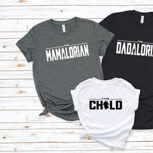 May include: Three t-shirts with white text on a light wood background. The shirts are gray, black, and white. The gray shirt says "The Mamalorian", the black shirt says "The Dadalorian", and the white shirt says "The Child".