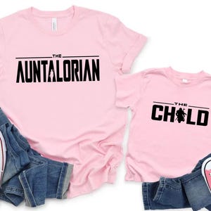 May include: Two light pink t-shirts with black text that reads "THE AUNTALORIAN" and "THE CHILD". The shirts are paired with blue jeans and white and pink sneakers. A fun matching set for family or friends.
