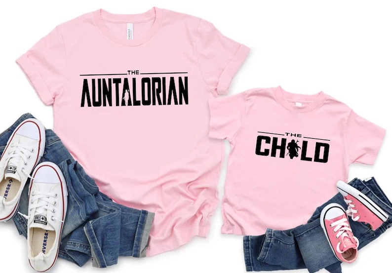 May include: Two light pink t-shirts with black text that reads "THE AUNTALORIAN" and "THE CHILD". The shirts are paired with blue jeans and white and pink sneakers. A fun matching set for family or friends.