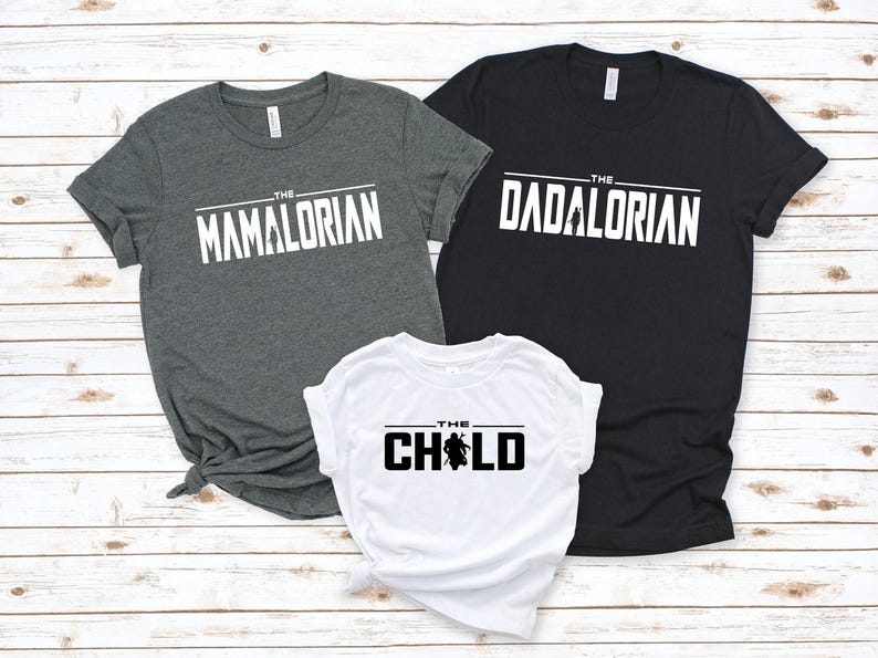 May include: Three t-shirts with white text on a light wood background. The shirts are gray, black, and white. The gray shirt says "The Mamalorian", the black shirt says "The Dadalorian", and the white shirt says "The Child".