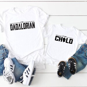 May include: White t-shirts with black text that reads "THE DADALORIAN" and "THE CHILD". The shirts are paired with blue jeans and white sneakers with black stripes, and black and white toddler shoes. A fun matching set.
