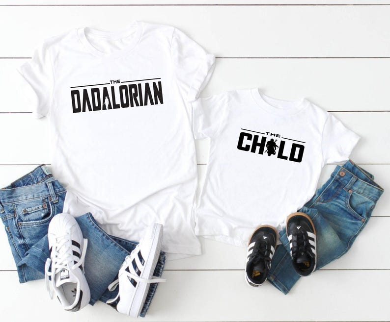 May include: White t-shirts with black text that reads "THE DADALORIAN" and "THE CHILD". The shirts are paired with blue jeans and white sneakers with black stripes, and black and white toddler shoes. A fun matching set.