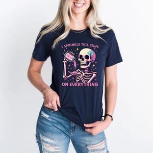 Puede incluir: Camiseta azul marino con un gr&aacute;fico de esqueleto colorido que sostiene un recipiente con la etiqueta "Sarcasmo". El texto de la camiseta dice "Yo esparzo esto sobre todo."