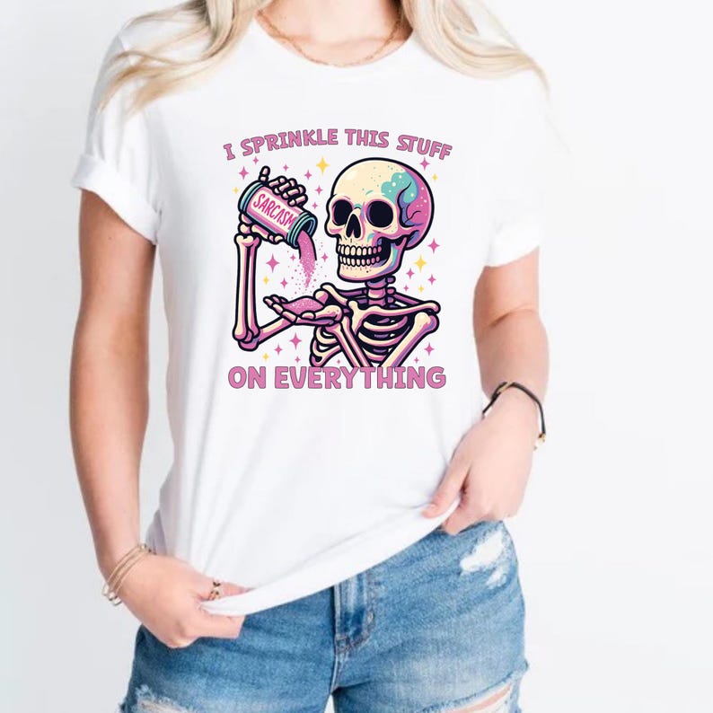 Puede incluir: Camiseta blanca con un gr&aacute;fico colorido de un esqueleto esparciendo "sarcasmo" de un recipiente. El texto "I sprinkle this stuff on everything" est&aacute; impreso encima y debajo de la ilustraci&oacute;n del esqueleto.