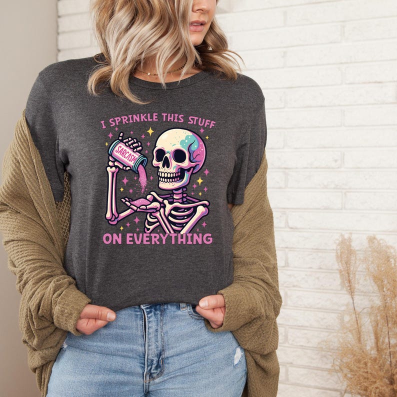 Puede incluir: Camiseta gris oscuro con un gr&aacute;fico de esqueleto colorido. El esqueleto sostiene un recipiente con la etiqueta "Sarcasmo" y lo est&aacute; rociando. El texto "I sprinkle this stuff on everything" est&aacute; impreso en la camiseta. El dise&ntilde;o incluye estrellas y colores pastel.