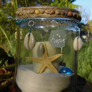 May include: A decorative glass jar with a beach theme. The jar contains sand, a starfish, a blue glass bead, and a candle. The lid is decorated with seashells and twine. The word "hope" is written on a piece of sea glass.