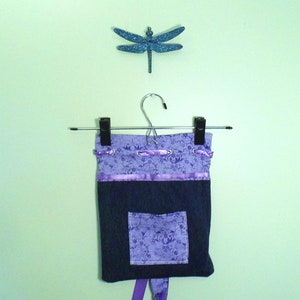 Könnte beinhalten: Eine handgefertigte Schürze mit einem dunkelblauen unteren Bereich und einem oberen Bereich mit lila Blumenmuster. Die Schürze hat eine Tasche und ist mit lila Band verziert. Über der Schürze befindet sich eine glitzernde blaue Libellen-Dekoration.