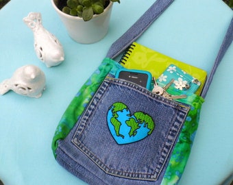 Può includere: Borsa a tracolla in denim con toppa a forma di cuore raffigurante la Terra. La borsa presenta una tasca, inserti in tessuto verde e blu e una tracolla in denim. All'interno sono visibili un taccuino, un telefono, un portafoglio e delle chiavi. Figurini decorativi in ceramica bianca e una pianta in vaso sono sulla superficie azzurra.