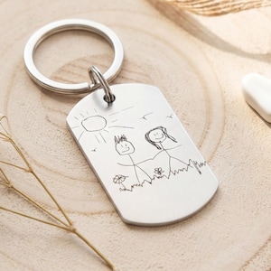 Op de afbeelding: Een zilverkleurige metalen sleutelhanger in de vorm van een dogtag met een zwarte gravure van twee stokfiguurtjes die onder een zon in een veld staan. Het rechthoekige plaatje heeft afgeronde hoeken en een ronde metalen ring.