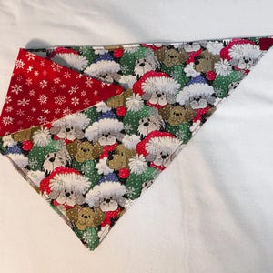 Pode incluir: Um bandana reversível para cão em vermelho e branco com uma estampa de Natal de cães usando chapéus de Papai Noel. Um lado apresenta um fundo vermelho com flocos de neve brancos, e o outro lado apresenta um fundo branco com uma estampa verde, azul e marrom de cães usando chapéus de Papai Noel.