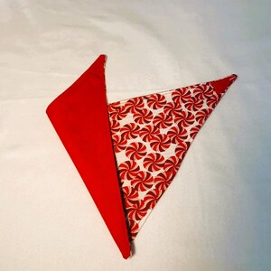 May include: A red and white bandana with a peppermint swirl pattern. The bandana is folded in half and the red side is facing out.