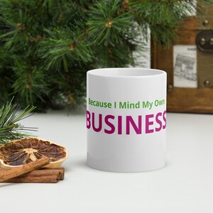 May include: White ceramic mug with the text "Because I Mind My Own BUSINESS" in green and magenta. The mug is set against a background of greenery and a wooden chest.