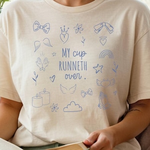 Peut inclure: T-shirt crème avec des gribouillis bleus et le texte "MY CUP RUNNETH OVER." Le t-shirt est associé à un short vert clair et des chaussettes blanches. La personne est assise sur un lit avec un livre dans les mains.