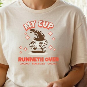 Peut inclure: T-shirt crème avec l'inscription "My Cup Runneth Over" et un dessin de tasse à café. La personne porte le t-shirt et est assise en tailleur, tenant un livre ouvert. L'ensemble dégage une atmosphère de détente.