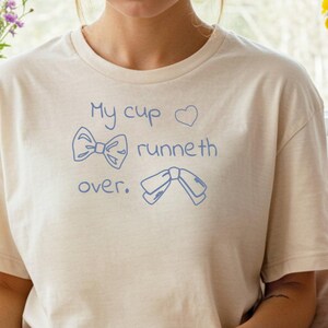 Peut inclure: T-shirt crème avec l'inscription bleue "My cup runneth over", orné de dessins de nœuds et de rubans. La personne porte un short vert clair et des chaussettes en tricot de câble blanches. Un livre ouvert est posé sur ses genoux.