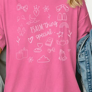 Peut inclure: Un t-shirt rose vif avec des illustrations blanches et le texte "PSALM thing special". Le motif comprend un cœur, une colombe, un nœud et d'autres symboles religieux. Le t-shirt a des manches courtes et un col rond.
