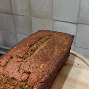 May include: A rectangular loaf of brown bread, freshly baked, rests on a wooden chopping board. The bread has a textured surface and a crack down the middle. The background shows a tiled wall.