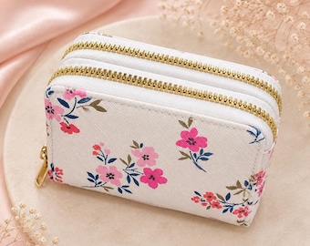 Può includere: Un portafoglio rettangolare bianco con un motivo floreale di fiori rosa e rossi e foglie verde scuro. Il portafoglio presenta due scomparti separati, ciascuno chiuso da una cerniera in metallo dorato con un cursore dorato abbinato.