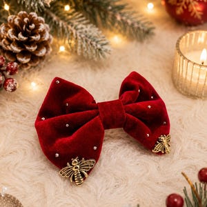 May include: A deep red velvet bow hair accessory featuring small embedded silver crystals and two gold-toned metal bee embellishments attached to the lower corners of the fabric, creating a structured and decorative hair piece.