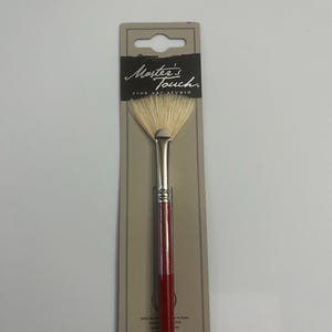 Op de afbeelding: Een Master's Touch Fine Art Studio waaiervormige kwast met een rode steel en witte haren. De kwast is verpakt in een bruine kartonnen achterkant met de merknaam erop gedrukt.